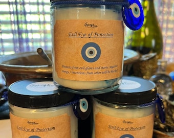 May include: Three glass jars of candles stacked on top of each other. Each jar has a black lid, a label with the text "Evil Eye of Protection", and a blue evil eye charm. The candles are white.