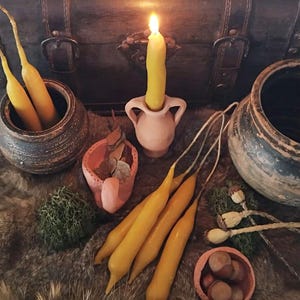 May include: A still-life arrangement featuring yellow taper candles, some lit, in small ceramic pots and holders. The scene includes a lit candle in a pink vase, a small bowl with nuts, and other decorative elements on a fur-like surface.