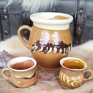 May include: A set of handmade ceramic mugs and a pitcher. The pitcher is a light brown with a cream-colored rim and a dark brown bear design. Two smaller mugs with handles are also included, featuring similar earth-tone colors and designs.