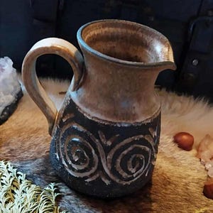 May include: A handmade ceramic pitcher with a dark brown base and a lighter brown top. The base features a carved spiral design. The pitcher has a curved handle and a spout for pouring. The pitcher is approximately 7 inches tall.