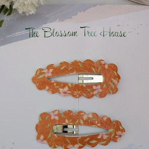 May include: Two orange hair clips with a floral pattern and scalloped edges. The clips have a gold metal bar.
