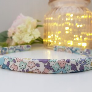 May include: A white headband with a floral pattern in shades of blue, purple, and pink. The headband is made of fabric and has a simple design.