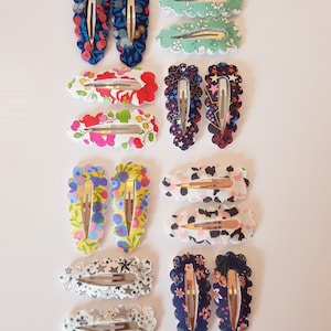 May include: Set of 12 hair clips with various floral and star patterns. The clips are made of fabric and have a scalloped edge. The clips are in pairs, with each pair featuring a different pattern.