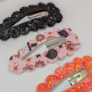 May include: Three Halloween-themed hair clips. One is black glitter, one is pink with a witch, cat, and candy corn print, and one is orange glitter.