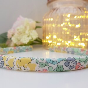 May include: A white headband with a floral print in shades of yellow, pink, blue, and green. The headband is made of fabric and has a simple design.