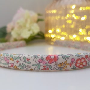 May include: A white headband with a floral print in shades of pink, green, and yellow. The headband is made of fabric and has a simple design. The letter "D" is visible in the bottom right corner of the image.