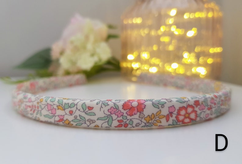 May include: A white headband with a floral print in shades of pink, green, and yellow. The headband is made of fabric and has a simple design. The letter "D" is visible in the bottom right corner of the image.