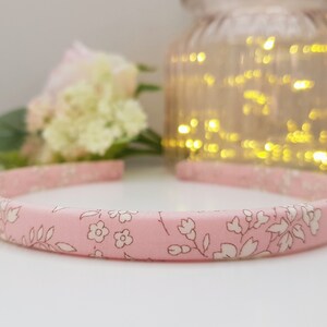 May include: Pink headband with a white floral pattern. The headband is made of fabric and has a simple design. The letter "E" is visible in the bottom right corner of the image.
