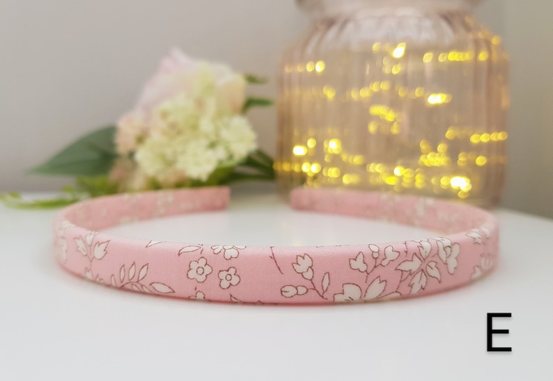 May include: Pink headband with a white floral pattern. The headband is made of fabric and has a simple design. The letter "E" is visible in the bottom right corner of the image.
