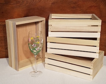 Peut inclure: Quatre caisses en bois empilées les unes sur les autres. Les caisses sont en bois naturel et ont une couleur brun clair. Un verre transparent avec un motif floral se trouve devant les caisses.