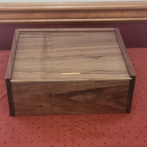 May include: A wooden box with a dark brown finish. The box has a hinged lid and a small, thin slot on the top.