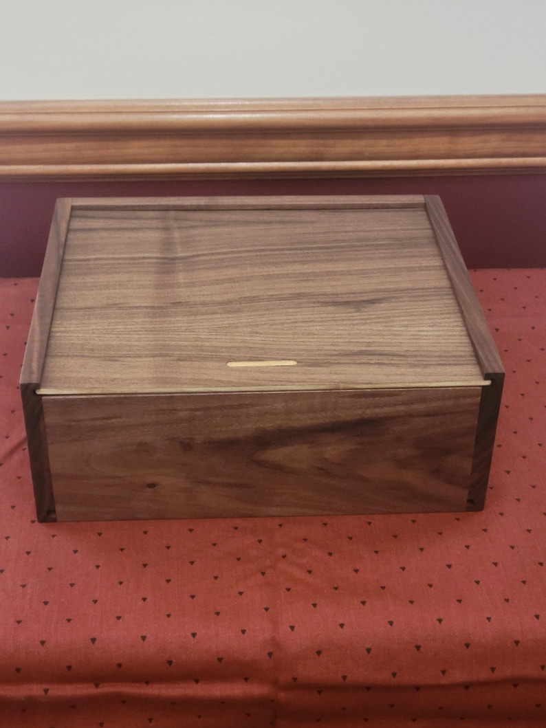 May include: A wooden box with a dark brown finish. The box has a hinged lid and a small, thin slot on the top.