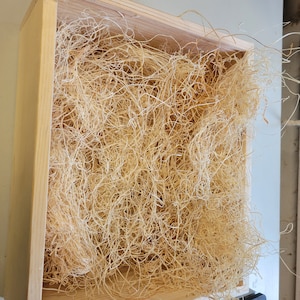 May include: A wooden box filled with light brown shredded paper, also known as packing peanuts, used for packaging and protecting fragile items.