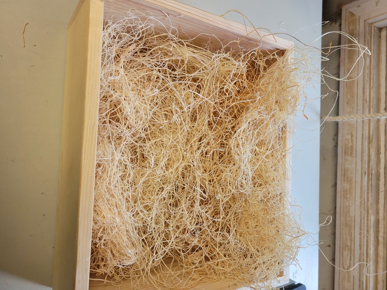 May include: A wooden box filled with light brown shredded paper, also known as packing peanuts, used for packaging and protecting fragile items.