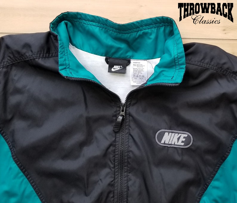 May include: Black and teal windbreaker jacket with a zipper closure and a Nike logo on the front. The jacket has a white tag with the Nike logo and size information.