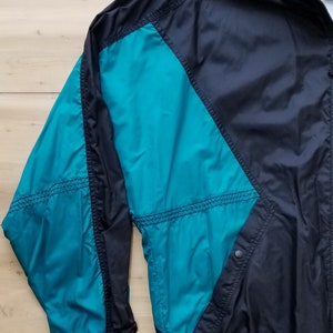 May include: A black and teal windbreaker jacket with a zipper closure and a drawstring at the waist. The jacket has a contrasting teal panel on the left sleeve and a teal collar.