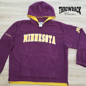 May include: Purple fleece hoodie with a yellow hood lining and a yellow "MINNESOTA" logo on the front. The hoodie has a yellow Columbia logo on the left sleeve and a yellow "M" logo on the right sleeve. The hoodie has a kangaroo pocket and a yellow drawstring.