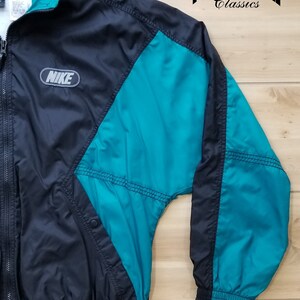 May include: A black and teal Nike windbreaker jacket with a zipper closure and a Nike logo on the left chest. The jacket has a teal panel on the right side and a teal panel on the left sleeve.