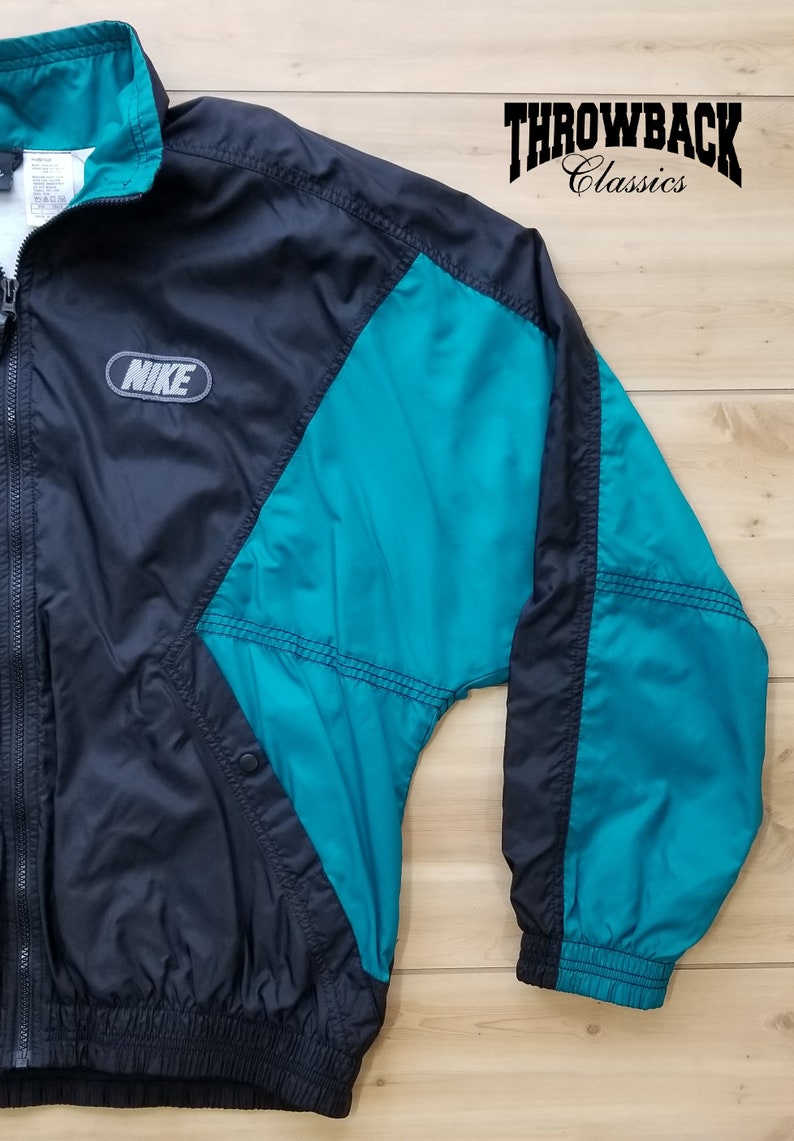 May include: A black and teal Nike windbreaker jacket with a zipper closure and a Nike logo on the left chest. The jacket has a teal panel on the right side and a teal panel on the left sleeve.