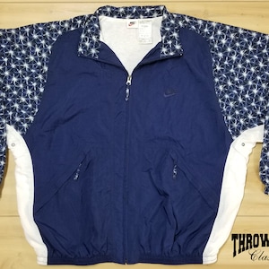 Op de afbeelding: Een donkerblauw windjack met witte accenten en een sterrenpatroon op de mouwen. Het jack heeft een ritssluiting en een Nike-logo op de borst.