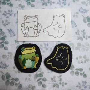 May include: Three embroidered patches, two frogs and a ghost. The frog patches are green and black with a stitched outline. The ghost patch is black with a yellow outline and a few small white stitches.