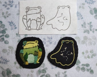 May include: Three embroidered patches, two frogs and a ghost. The frog patches are green and black with a stitched outline. The ghost patch is black with a yellow outline and a few small white stitches.