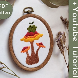 May include: Embroidered hoop art featuring a mushroom with a green frog sitting on top. The mushroom has red, orange, and yellow stripes. The hoop is made of wood and has a brown stain.