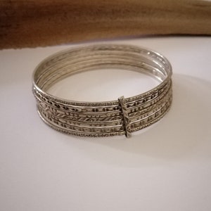 Puede incluir: Un conjunto de pulseras de plata con intrincados diseños grabados. Las pulseras están conectadas por un cierre de plata. Las joyas se exhiben sobre una superficie blanca con un objeto marrón borroso en el fondo.