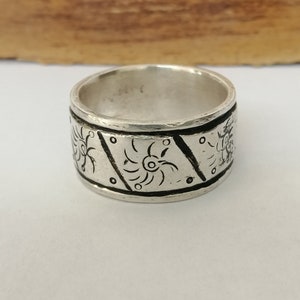 May include: A silver ring with a wide band featuring an intricate design of repeating black floral patterns.