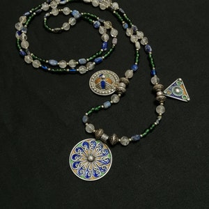 May include: A long silver necklace with blue, green, and white beads. The necklace has two pendants, one round and one triangular, both with intricate designs and blue, green, and orange enamel.