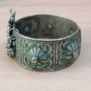 May include: A silver bracelet with intricate floral designs and a chain clasp. The bracelet has a green patina and is decorated with raised, circular accents.