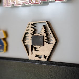 Op de afbeelding: Een zeshoekige houten magneet met een uitgesneden ontwerp van twee dennenbomen. De magneet is lichtbruin van kleur en heeft een zwarte magnetische achterkant.