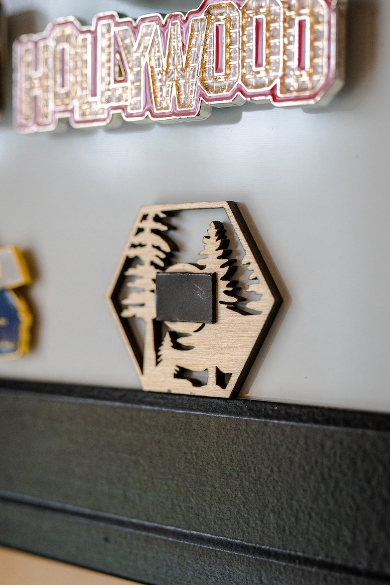 Op de afbeelding: Een zeshoekige houten magneet met een uitgesneden ontwerp van twee dennenbomen. De magneet is lichtbruin van kleur en heeft een zwarte magnetische achterkant.