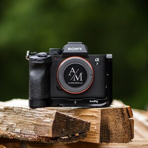 May include: A black Sony α7 camera with a red-ringed lens is displayed on a stack of wood. The lens features a circular logo with the initials "A/M" and the text "ALLISON MEDIA CO." The camera is labeled "SONY" and "α 7".