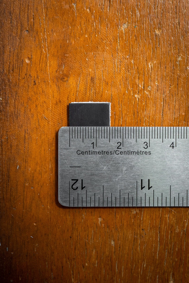 Op de afbeelding: Een close-up van een metalen liniaal met zwarte markeringen. De liniaal is gemarkeerd in centimeters en inches. De liniaal meet een klein zwart vierkant.