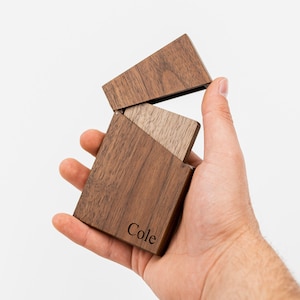 May include: A rectangular wooden box with a hinged lid, held open. The box is made of dark brown wood and has the name "Cole" engraved on the front. The box has a smooth finish and is held in a hand.