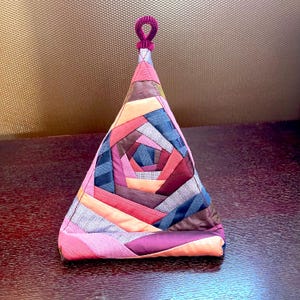 May include: A colorful, geometric-patterned tea cozy. The cozy is shaped like a pyramid, with a spiral design in shades of pink, purple, orange, and blue. A small, looped handle is at the top. The cozy is made of fabric.
