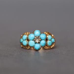 Puede incluir: Un anillo de oro con un diseño floral con piedras turquesas y un diamante central. El anillo tiene una banda detallada y texturizada y un grupo de piedras turquesas azul brillante dispuestas en forma de flor.