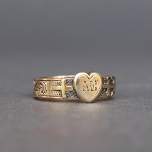 May include: A gold-toned ring with a heart-shaped centerpiece. The heart features engraved letters. The band has decorative patterns and cross-like designs. The ring is set against a neutral gray background.