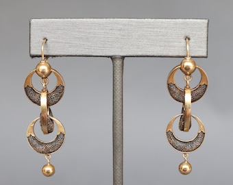 May include: A pair of gold tone earrings with a dangling design. Each earring features two circular hoops with a brown mesh fabric design and a small gold ball at the bottom.
