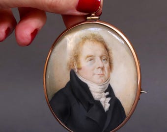 May include: An antique oval portrait miniature in a gold-tone frame. The portrait depicts a man with fair skin, light hair, and a black coat. The miniature is held by a hand with red nail polish.