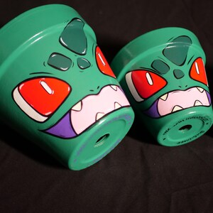 Peut inclure: Des pots de fleurs en céramique verte avec un motif de visage de monstre de dessin animé. Les visages ont des yeux rouges, des dents blanches et des accents violets. Les pots sont peints avec une finition brillante.