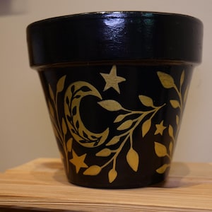 Puede incluir: Maceta de terracota pintada de negro con un diseño celestial dorado con una luna creciente, estrellas y vides con hojas. La maceta está sobre una superficie de madera.