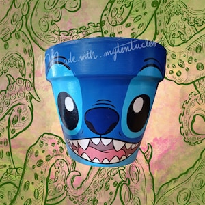 Op de afbeelding: Een blauwe en witte keramische plantenpot beschilderd met een cartoonfiguur dat lijkt op Stitch uit de Disney-film Lilo & Stitch. De pot heeft een brede opening en een ronde bodem. De achtergrond is een groen en roze aquarelontwerp met een patroon van tentakels.