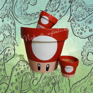 Pode incluir: Três vasos de flores vermelhos e brancos com tema de cogumelos com rostos de desenhos animados. Os vasos são pintados com uma base vermelha, detalhes brancos e olhos pretos. O fundo apresenta tentáculos de polvo verdes e verde-azuis.