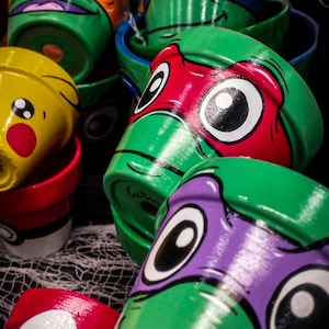 Può includere: Una collezione di vasi di fiori in ceramica dipinti a mano con personaggi dei cartoni animati popolari. I vasi sono dipinti con colori vivaci, tra cui verde, arancione, blu, rosso, giallo e viola. I disegni includono le Tartarughe Ninja e un personaggio Pokémon.