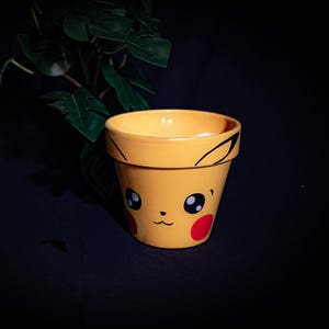 Op de afbeelding: Gele keramische bloempot met een Pikachu-ontwerp. De pot heeft zwarte oren, ogen en een mond, met rode wangen. De pot is ontworpen voor kleine planten en is een decoratief item.
