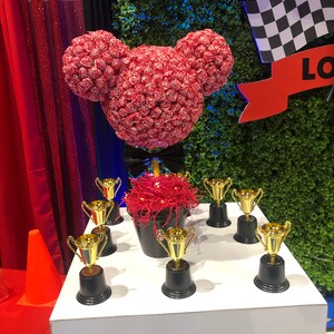 Könnte beinhalten: Ein roter und weißer Mickey-Maus-Kopf aus Süßigkeiten steht auf einem Tisch mit einer schwarzen Fliege. Auf dem Tisch stehen acht goldene Pokale.