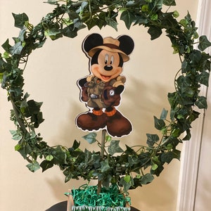 Könnte beinhalten: Ein grüner Efeu-Kranz mit einem ausgeschnittenen Mickey-Maus-Charakter in der Mitte. Der Charakter trägt einen Safarihut und hält einen Kompass. Der Kranz sitzt auf einer kleinen braunen und grünen Box mit grünem grasähnlichem Material im Inneren.