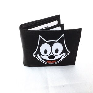Könnte beinhalten: Schwarze Ledergeldbörse mit einem Comic-Katzenkopf auf der Vorderseite. Die Katze hat große weiße Augen und ein breites Lächeln. Die Geldbörse hat einen Kreditkarten-Steckplatz.